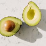 Two avocado halves on a white marble surface, one holding the large brown pit