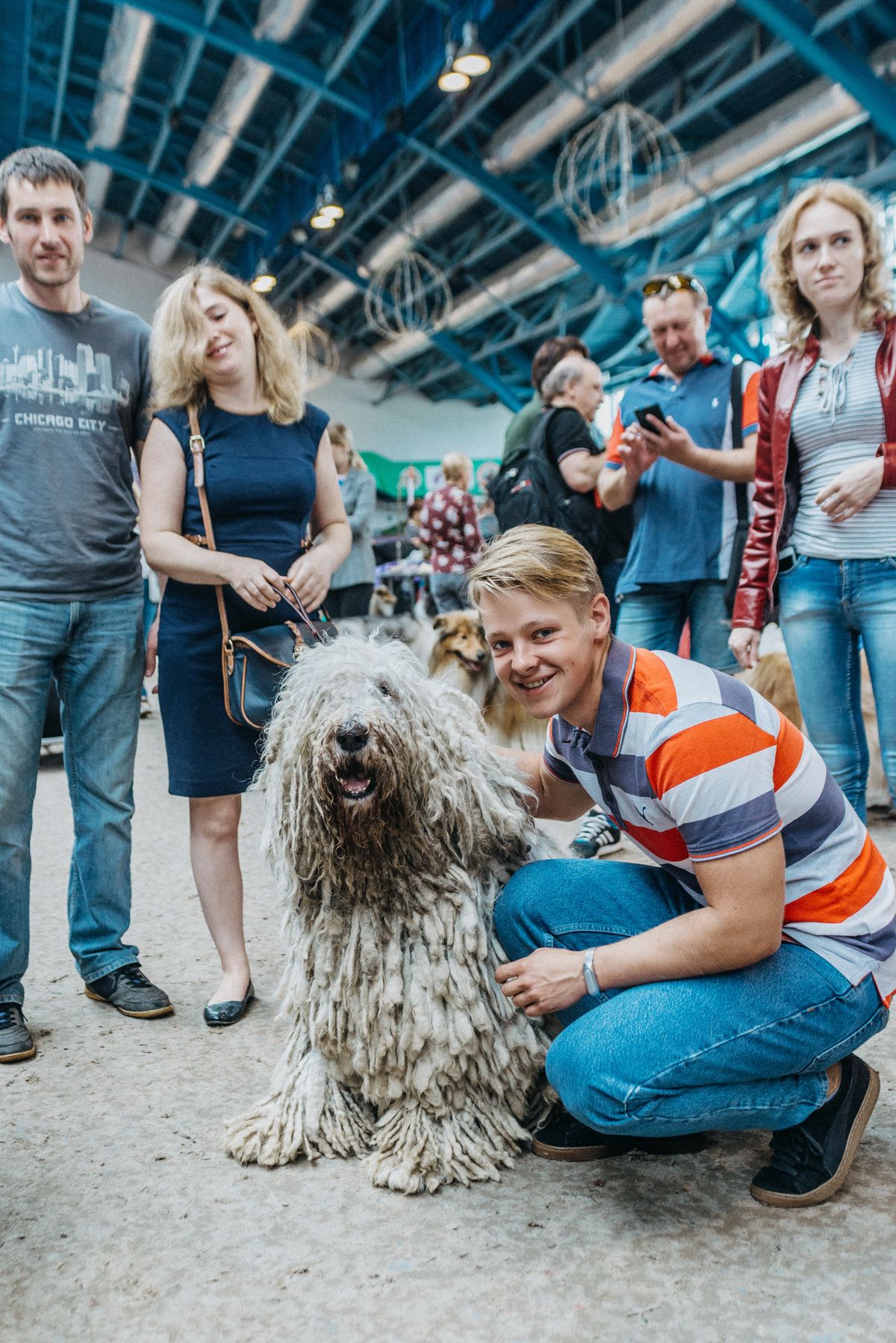 Smiling visitor crouching beside a corded Komondor dog at a busy indoor dog show